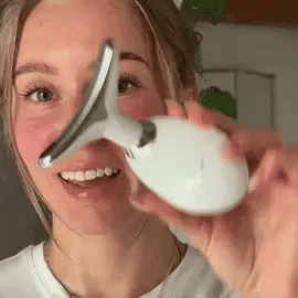 Animated GIF of a smiling young woman demonstrating how to hold the SkinRevive LED face massager against her cheek for a daily lifting skincare routine.
